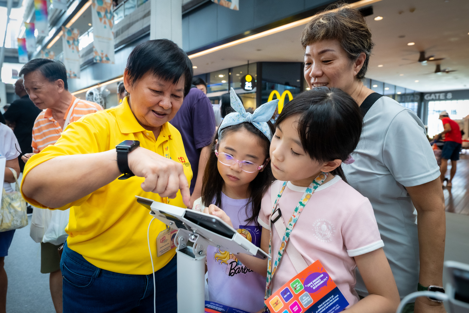 Person in yellow shirt points at a tablet on a stand as two children and an adult watch in a busy hall.